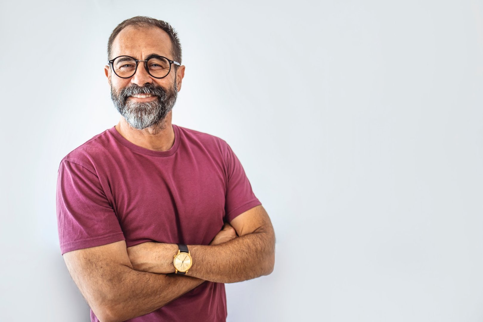Portrait of happy mature man wearing spectacles and looking at camera indoor. Man with beard and glasses feeling confident.  Handsome mature man posing against a grey background